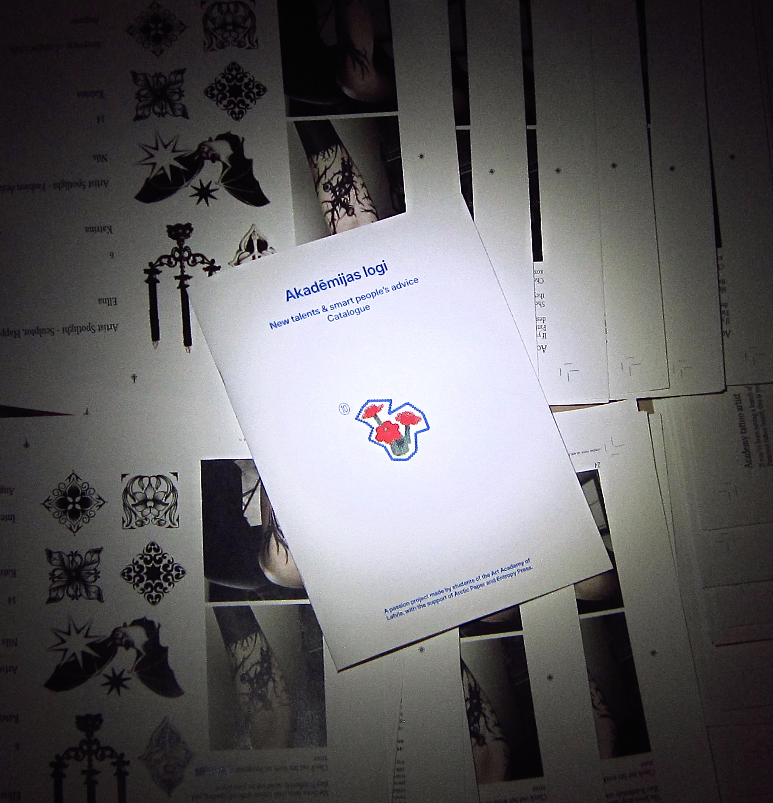 Akadēmijas logi magazine resting on printed artwork and catalogue pages.